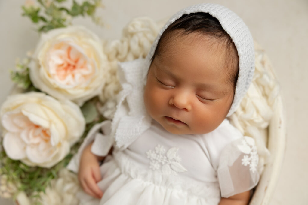 Close-up portrait of newborn Emmy wearing a white bonnet surrounded by cream florals during her newborn photography session in Fresno, California.